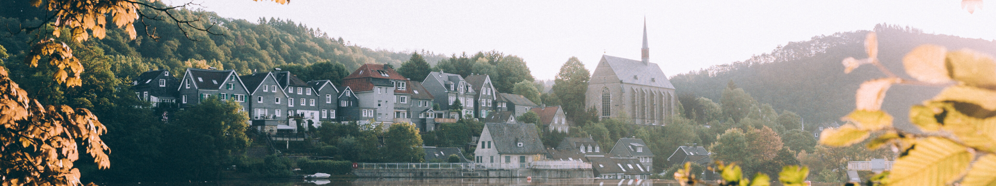 Blick durch die Blätter über den Beyenburger Stausee, in dem sich die Fachwerkhäuser vom anderen Ufer spiegeln.  