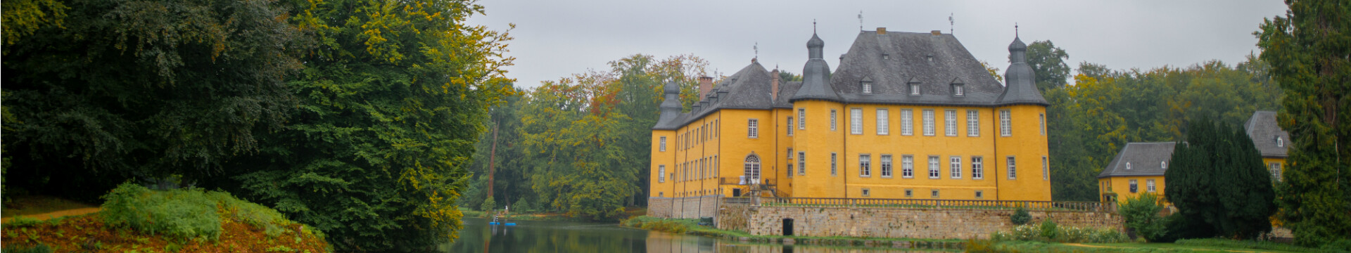 Blick auf das historische Schloss Dyck in Jüchen am Niederrhein, umgeben von einem weitläufigen Schlosspark. Das gelb gestrichene Wasserschloss mit seinen markanten Türmen spiegelt sich im ruhigen Wasser des Schlossgrabens wider. Im Vordergrund sind gepflegte Rasenflächen und Wege zu sehen, die von üppiger Vegetation und hohen Bäumen umgeben sind.