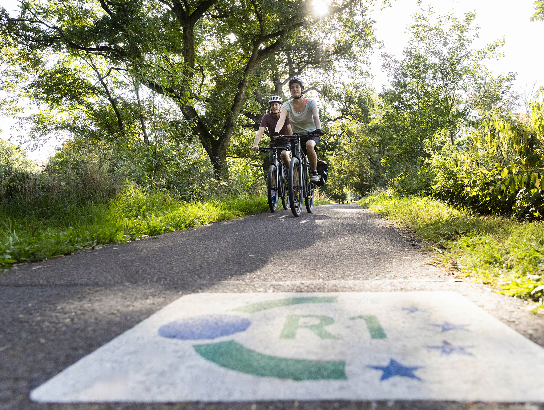 Radfahrende auf dem über den Europaradweg R1. Auf dem Boden ist das Logo des R1 zu sehen. 