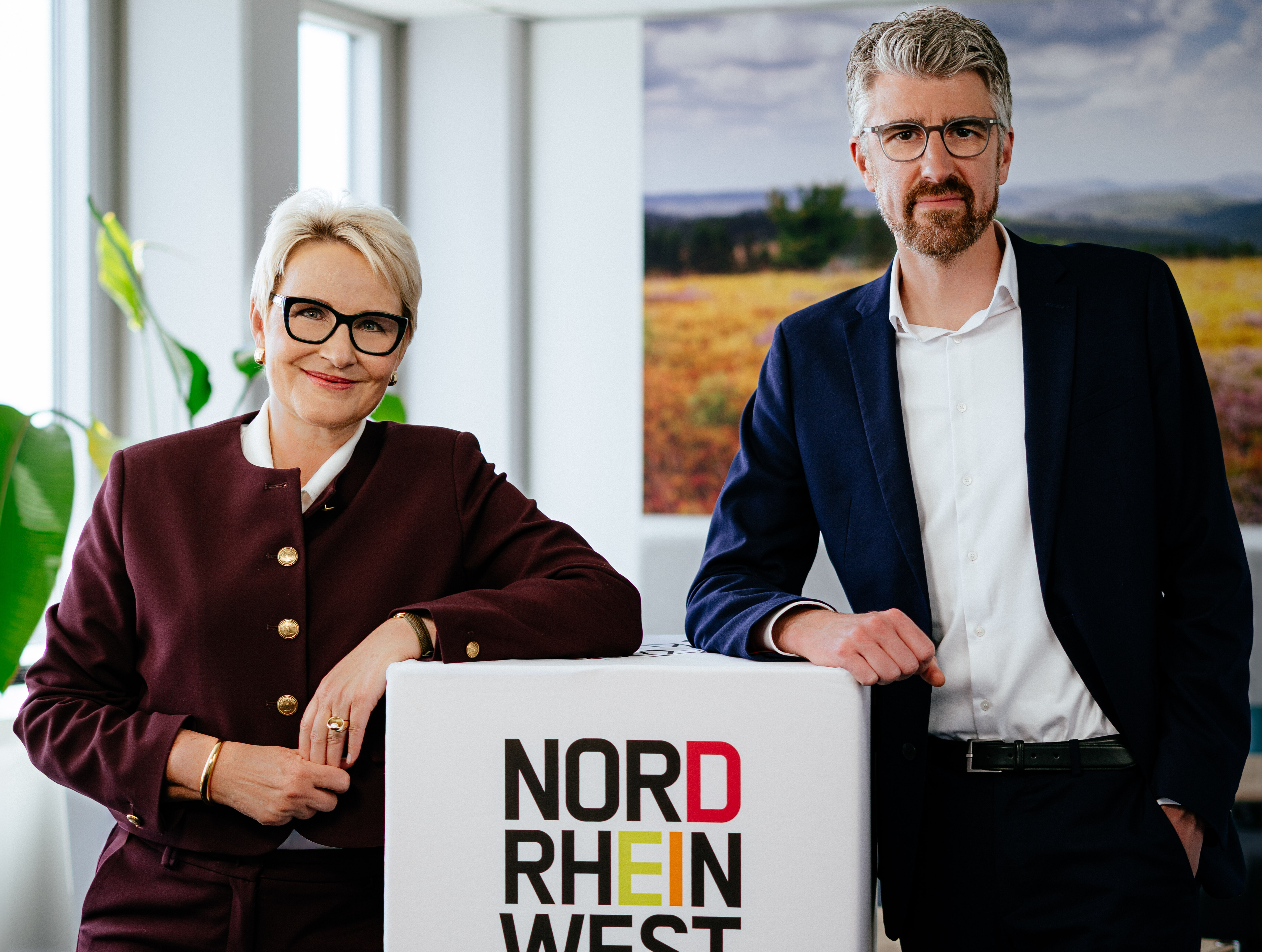 Die Vorstandsvorsitzende von Tourismus NRW, Dr. Heike Döll-König, und ihr Stellvertreter, Sebastian Kaiser, stehen in einem Konferenzraum neben einem großen Würfel, auf dem das Logo des Reiselands Nordrhein-Westfalen zu sehen ist. 
