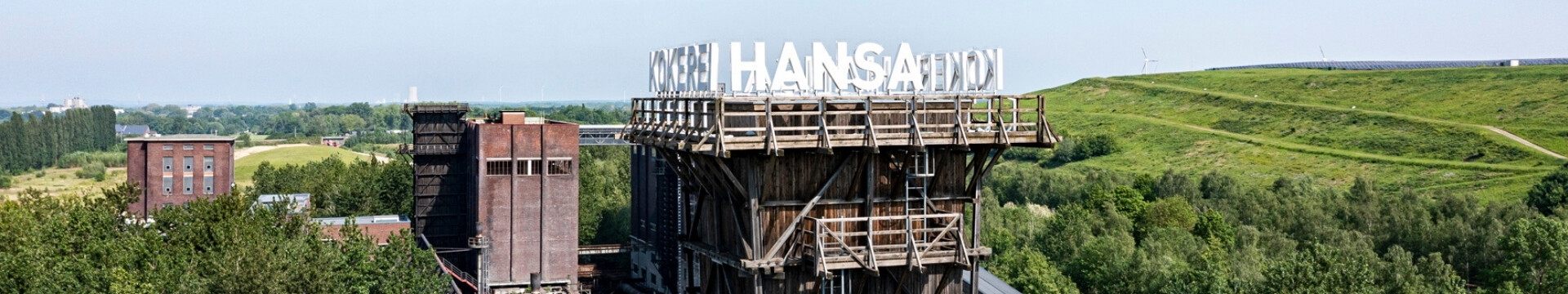Turm der Kokerei Hansa in Dortmund, umgeben von Bäumen und dem grünen Deusenberg, einem rekultivierten Deponiegelände. Über dem Bild stehen die Logos von Tourismus NRW und Ruhr Tourismus. 