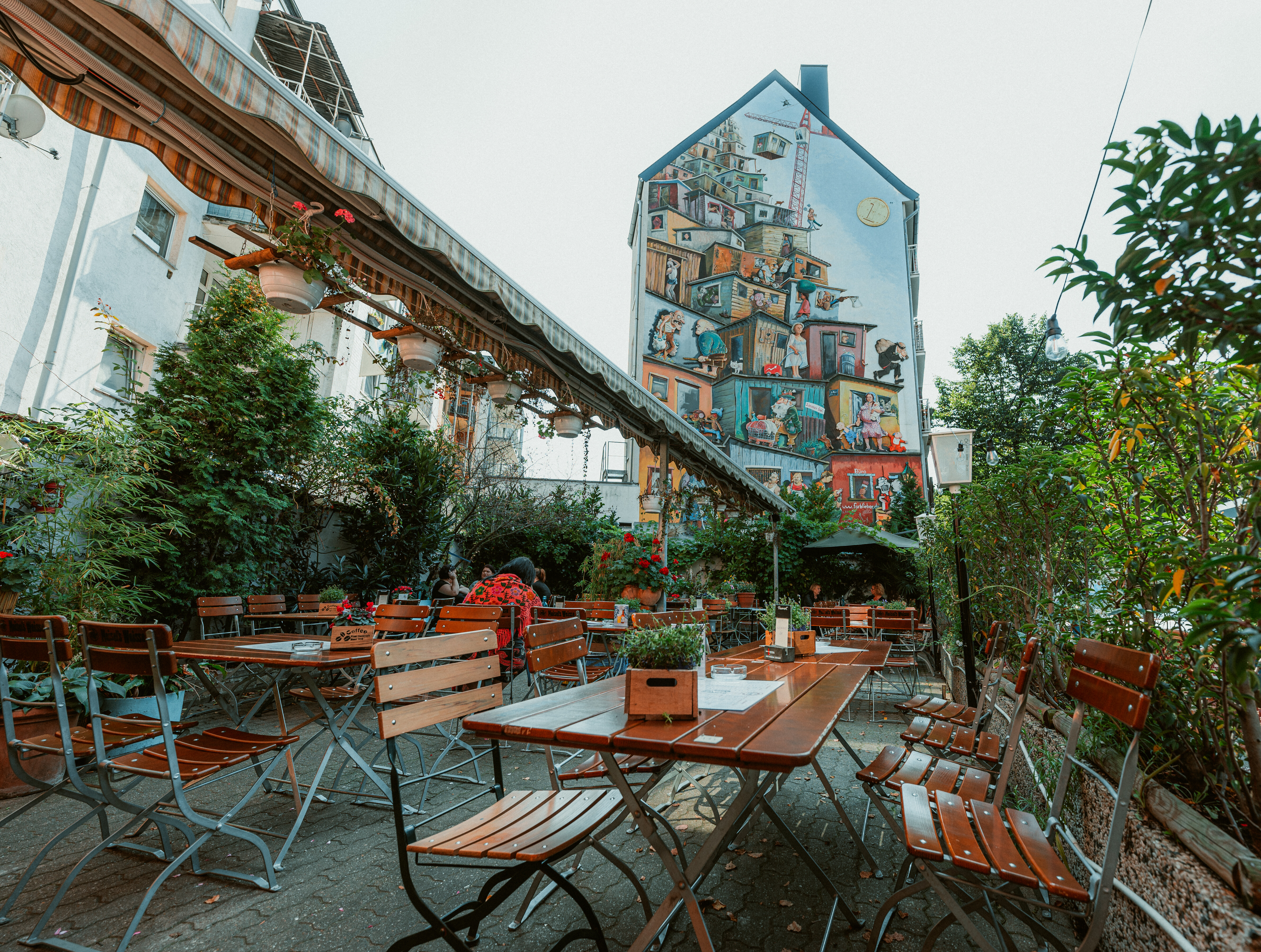 Tische und Stühle vor in einem Lokal in der Brunnenstraße in Düsseldorf. Im Hintergrund ist ein großes Mural an einem Haus zu sehen.  