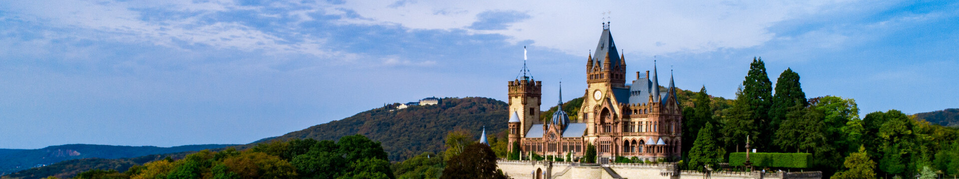 Schloss Drachenburg steht bei Königswinter auf einem Hügel, umgeben von dichtem Wald. Die Burg hat mehrere Türme mit spitzen Dächern. Um die Burg erstrecken sich bewaldete Hügel. Der Himmel ist blau mit leichten Wolken.