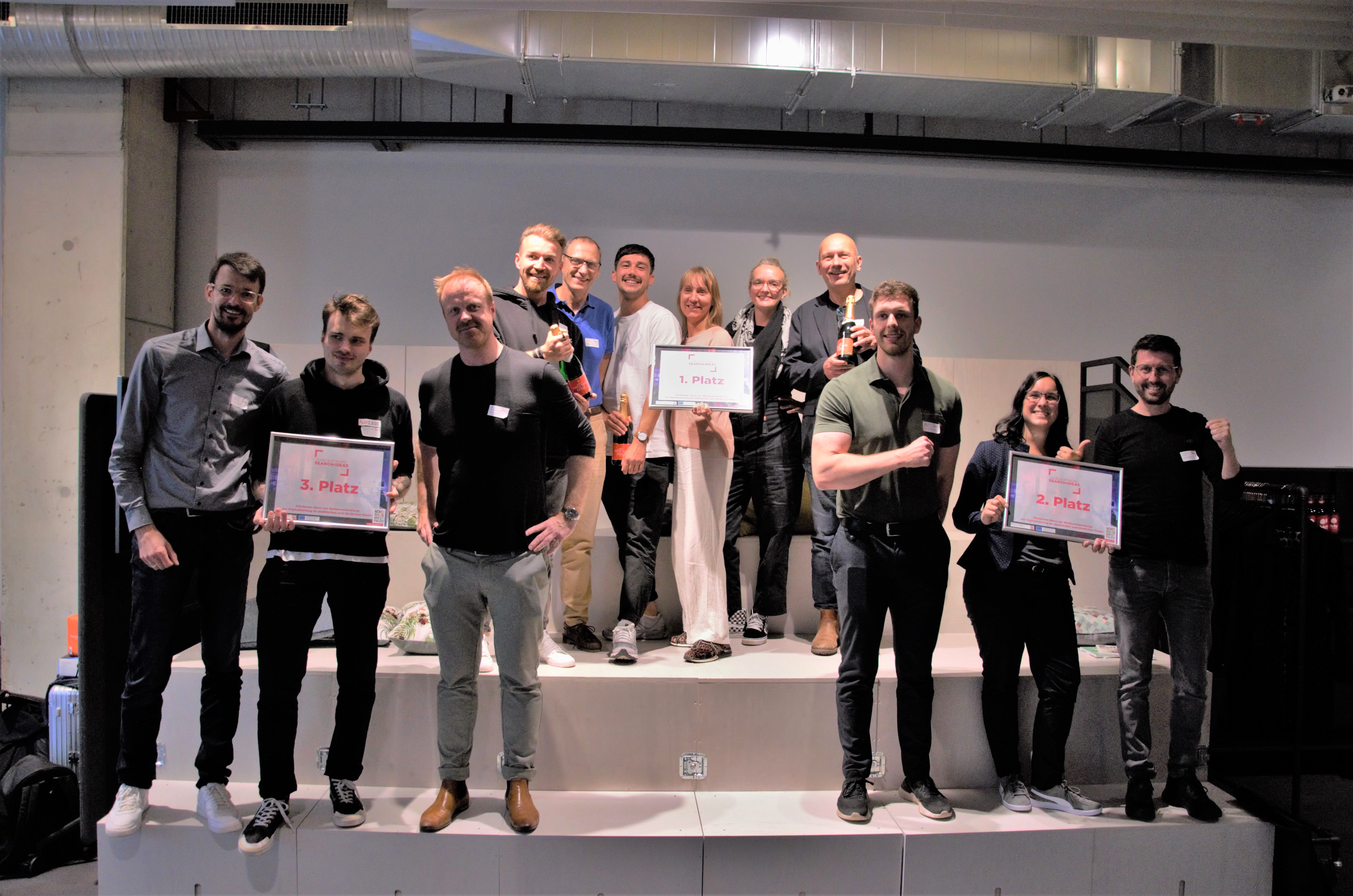 Drei Gruppen von Menschen halten Schilder mit "1. Platz", "2. Platz" und "3. Platz" hoch. Es handelt sich dabei um die Teams mit den besten Ideen beim 1. Hackathon des Tourismus NRW. 