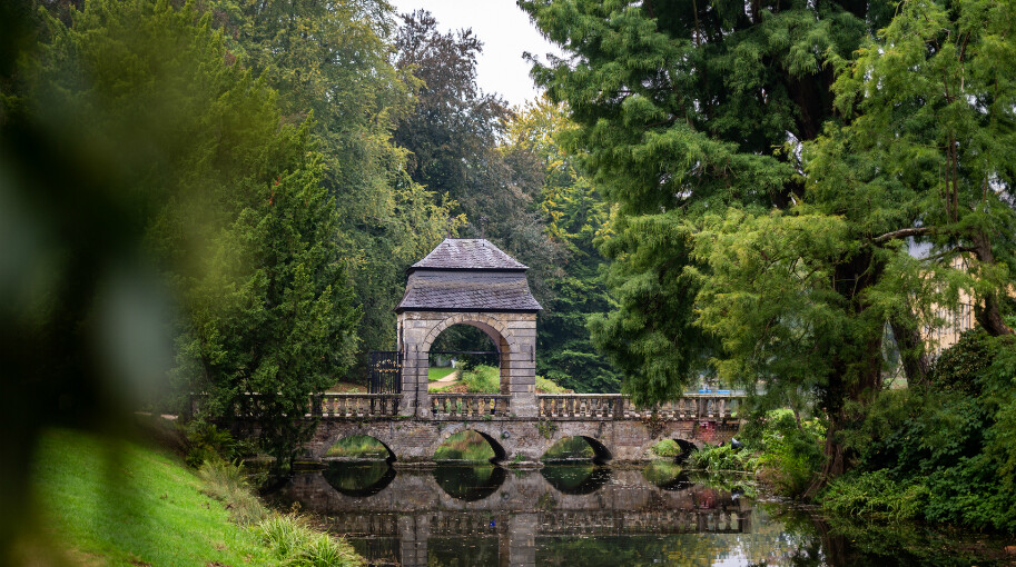 Ein steinernes Torhaus steht im Park von Schloss Dyck in Jüchen am Niederrhein auf einer Brücke, die ein ruhiges Gewässer überquert. Die Brücke hat mehrere Bögen, die sich im Wasser spiegeln. Das Torhaus ist von dichtem Grün umgeben, darunter große Bäume und Büsche. 