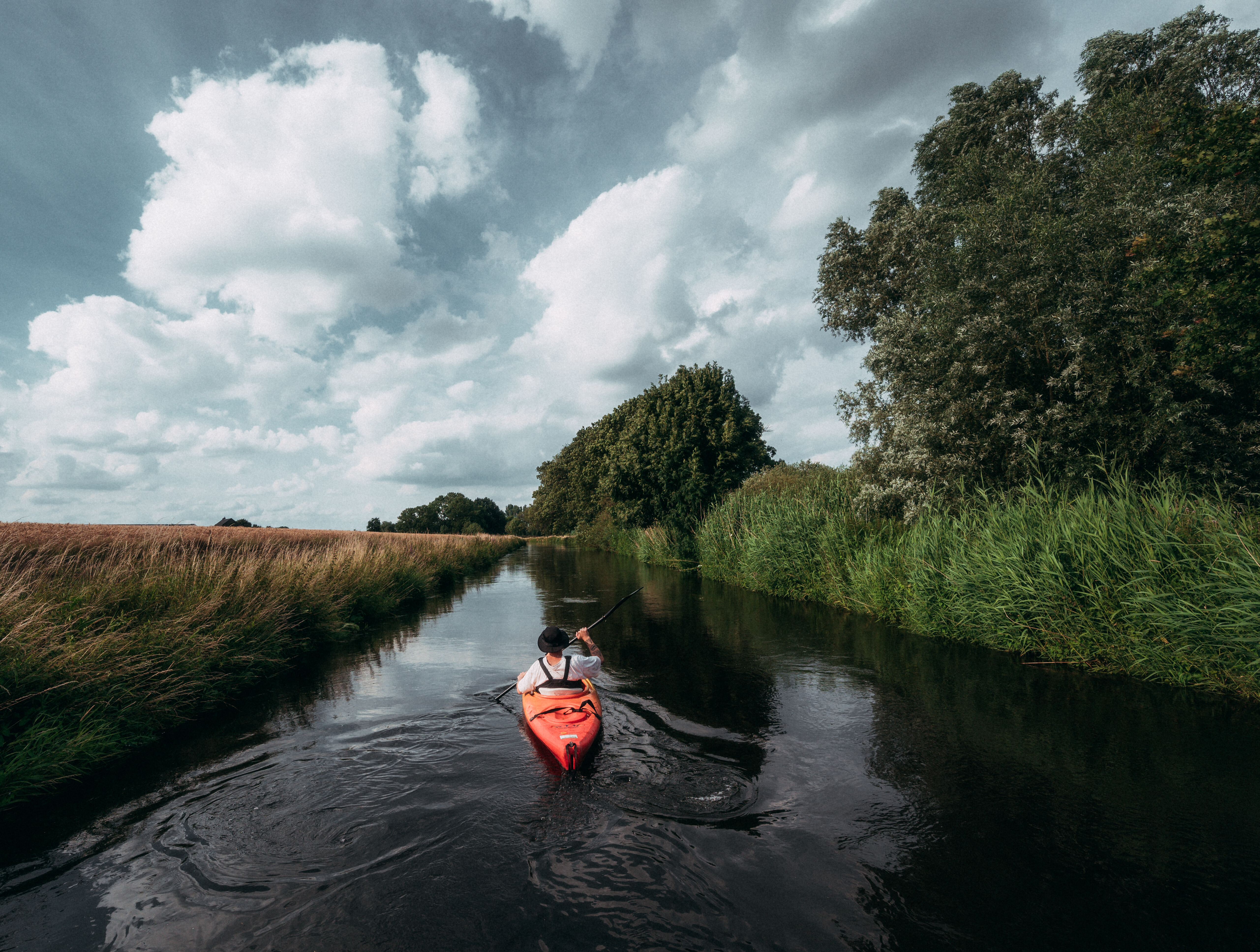 Ein Mensch fährt mit einem roten Kajak fahren auf der Niers. Die Flussauen sind dicht mit Gräsern und Bäumen bewachsen. 