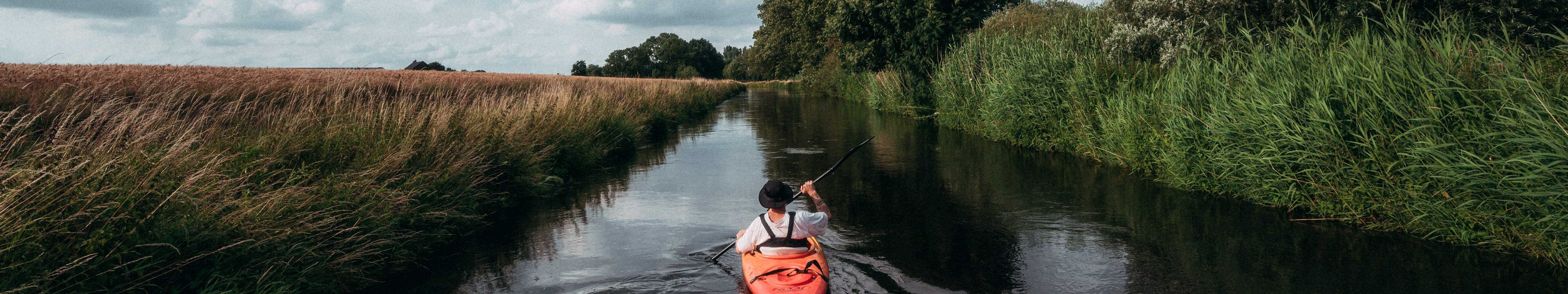 Ein Mensch fährt mit einem roten Kajak fahren auf der Niers am Niederrhein. Die Flussauen sind dicht mit Gräsern und Bäumen bewachsen.  © Tourismus NRW e.V./Johannes Höhn