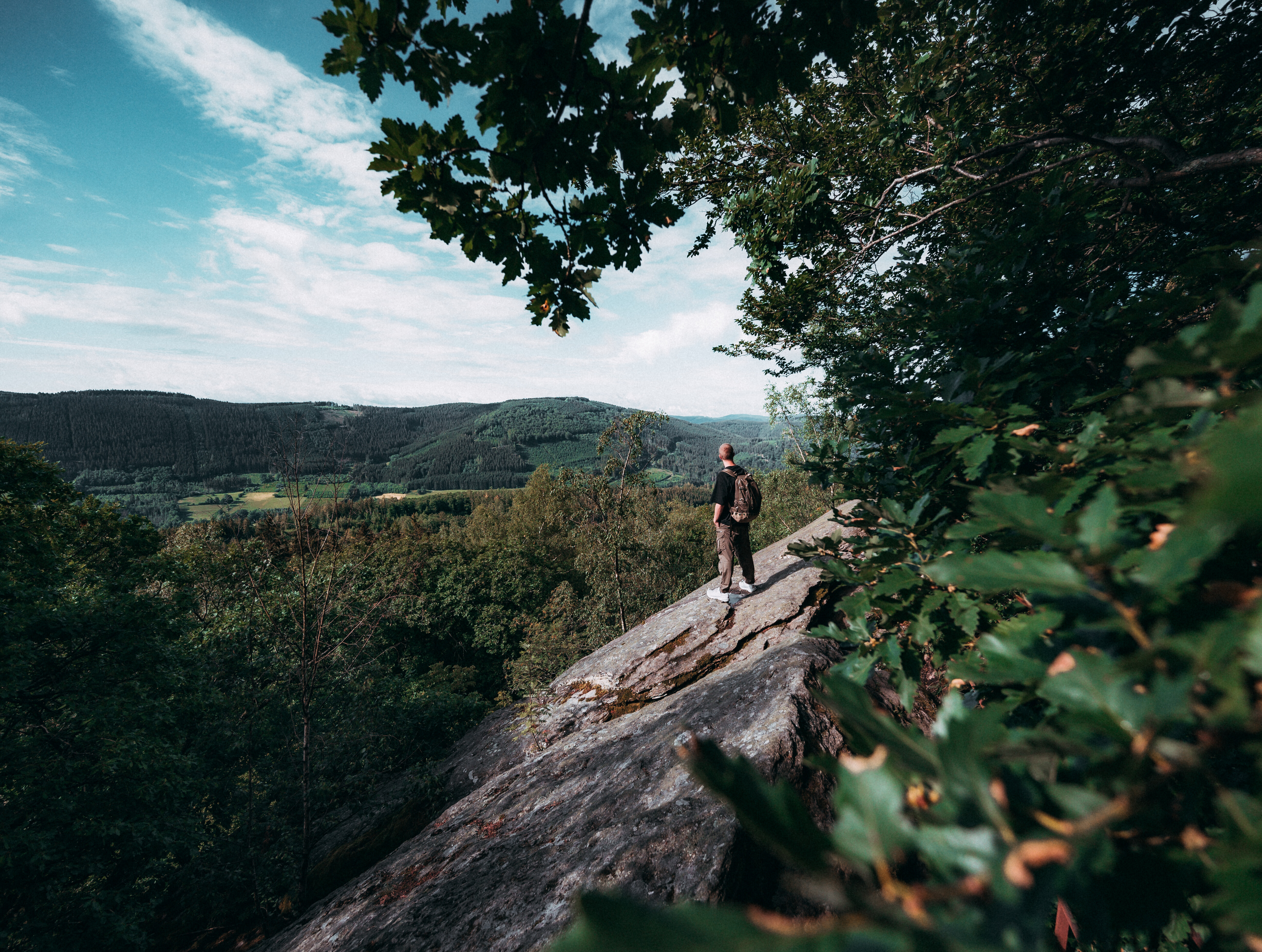 Ein Wanderer steht auf einem Felsvorsprung am Walderlebnispfad Saalhausen im Sauerland und blickt über die bewaldeten Hügel. 