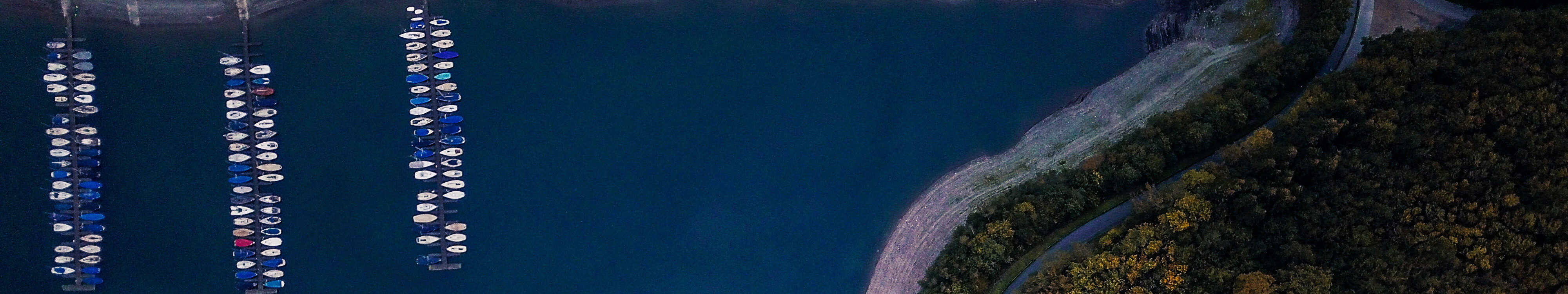 Der Biggesee und ein angrenzendes Waldstück aus der Vogelperspektive. Drei Stege mit kleinen Booten ragen in den tiefblauen See. 
