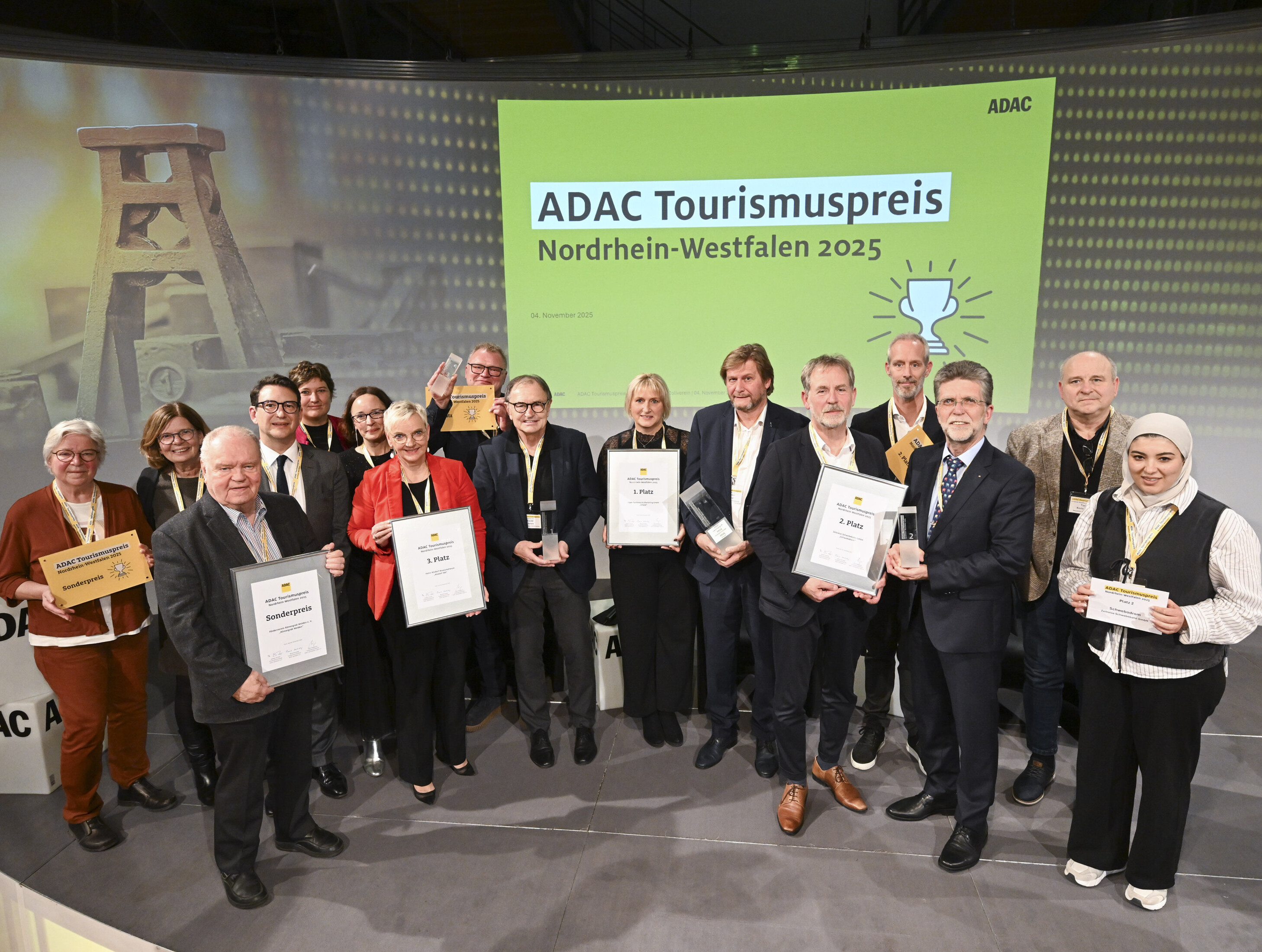 Gewinner des ADAC-Tourismuspreises NRW 2025 © Andreas Buck Die Gewinner des ADAC-Tourismuspreises NRW 2025 stehen im Halbkreis und halten ihre Urkunden in der Hand. Hinter sie sind Bilder von Zeche Zollverein und dem Logo des Tourismuspreises projiziert.