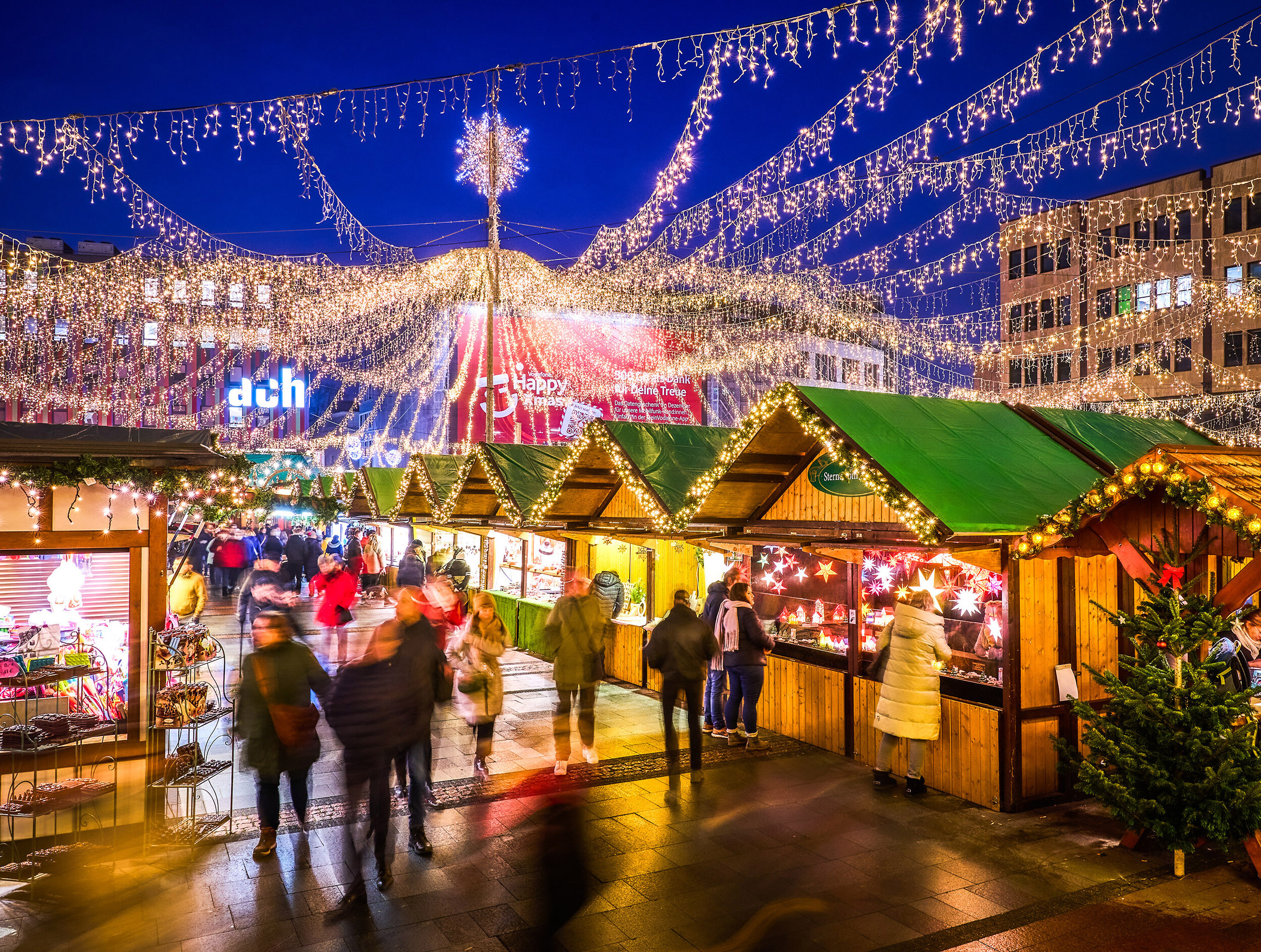 Festlich beleuchteter Internationaler Weihnachtsmarkt in Essen am Abend. Über den Holzhütten sind Lichterketten gespannt, die sich strahlenförmig von einem zentralen Punkt mit einem leuchtenden Stern aus nach außen verteilen und den Markt in ein warmes, gemütliches Licht tauchen. Besucher schlendern durch die Gänge zwischen den Buden, die verschiedene Produkte anbieten. 