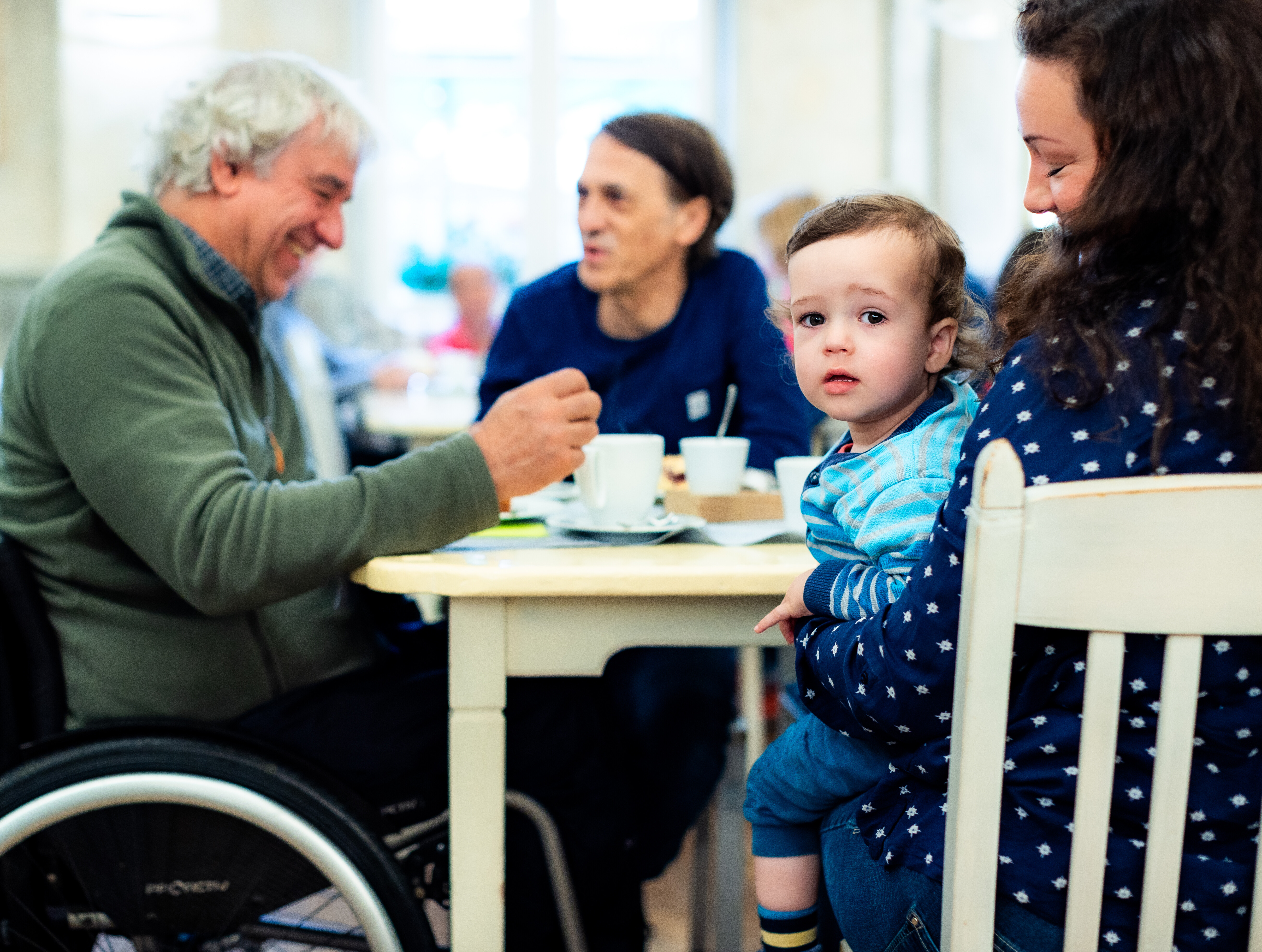 Älterer Mann mit grauen Haaren im Rollstuhl lacht und unterhält sich mit einem Mann mittleren Alters an einem Café-Tisch. Eine Frau mit braunen Locken hält ein Kleinkind auf dem Schoß; das Kind blickt neugierig in die Kamera. Auf dem Tisch stehen Kaffeetassen und Unterteller. 
