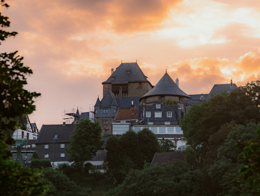 Schloss Burg in Solingen, vor einem Himmel mit orange leuchtenden Wolken bei Sonnenuntergang, umgeben von Häusern und Bäumen.