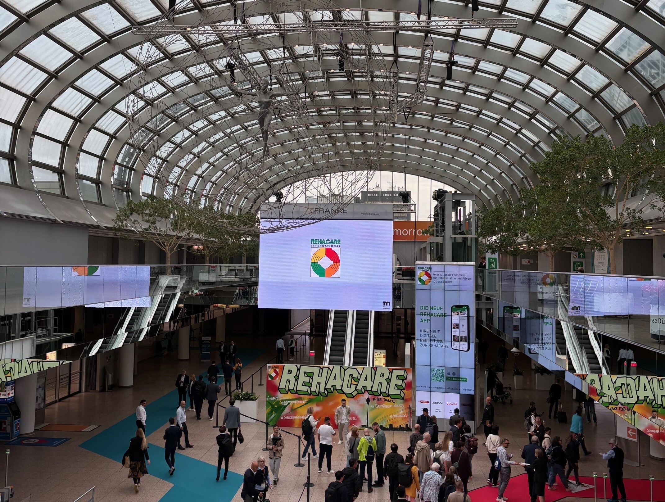 Blick in den Eingangsbereich der Messe Essen mit einer gläsernen Kuppel. Viele Menschen sind unterwegs, mehrere Schilder verweisen auf die Rehacare 2025.  